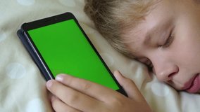 Closeup view of cute funny kid pretending to be sleeping in bed in morning. Young boy holds smartphone with blank greenscreen in hand. Real time 4k video footage.