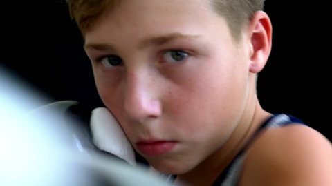 The boxer boy stands on a dark background, the boy has a gray T-shirt, his face is almost covered in gloved hands. The boy has light short hair and dark eyes.
