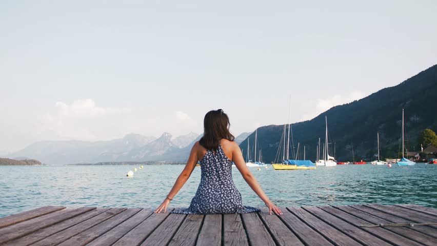Young woman in dress sitting at the edge of wooden pier at lake in mountains, enjoying view