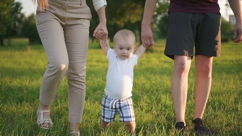 Lovely family teaches infant to first walk in sunny park. Mother and father holding hands of little son and moving on the green ground. Outdoors.