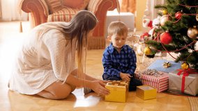 4k video of adorable boy sitting on floor with mother under Christmas tree and opening gift box with presents