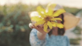 Close up girl in hat twists flower of sunflower on extended hand in front of camera , cheerful young woman, countryside style, lifestyle slow motion video shooting by handheld gimbal