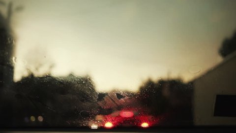 Slow motion rain flows and wipers on the front windshield glass window of car on the road with beautiful colorful blurry light of traffic outside street vehicle city raindrop travel weather close up
