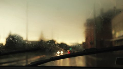 Close up rain flows and wipers on the front windshield glass window of car on the road with beautiful colorful blurry light of traffic outside street vehicle city raindrop travel weather slow motion