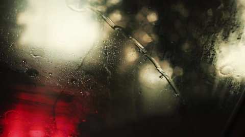 Close up rain flows and wipers on the front windshield glass window of car on the road with beautiful colorful blurry light of traffic outside street vehicle city raindrop travel weather slow motion