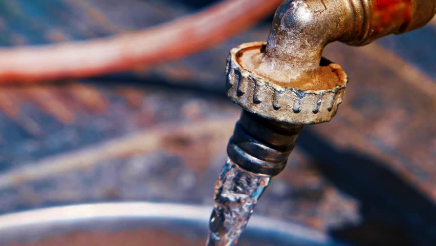Resource conservation: dramatic macro capture of antique brass faucet with oxidized patina, showcasing crystalline water droplet clinging to spout tip under brilliant daylight. Intricate threading patterns reveal metallic corrosion and weathering across amber, golden, and earthy brown tones that suggest advanced age and industrial neglect. Single luminous bead of water catches sunlight with prismatic clarity, forming spherical jewel-like focal point that reflects microscopic detail and ambient illumination. Shallow depth of field renders background elements in soft focus with blurred bokeh effects, revealing muted blues, grays, and rust-red hues of additional corroded piping. Natural overhead lighting casts subtle shadows across mineral deposits and surface irregularities, emphasizing three-dimensional form and material degradation. Tightly framed composition transforms utilitarian object into compelling study of environmental awareness, infrastructure decay, and visual metaphor for precious natural resources.