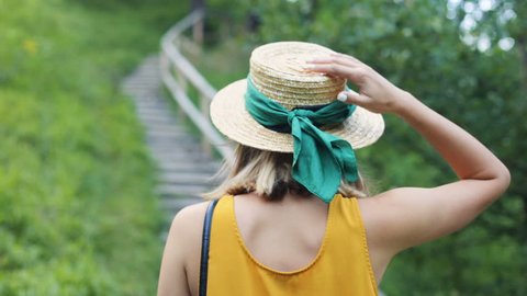 Nice young Caucasian woman wearing yellow t-shirt walking up the stairs. Slavic girl touching hat. Outdoors. Summertime. Back.