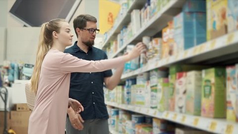 Married couple is buying baby formula in supermarket. They are standing near shelf and taking different jars from it, reading ingredients