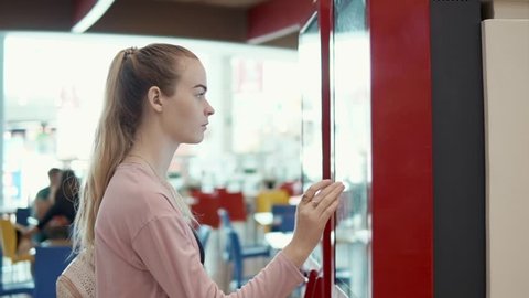 Young blonde girl ordering food in fast food restaurant using self-service payment machine, self-service kiosk. Consumer using technologies to buy food.
