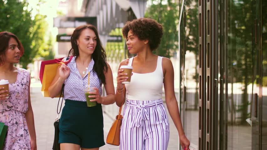 sale, consumerism and people concept - happy young women with shopping bags and drinks walking along city street