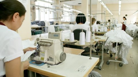 Female workers using sewing machines while working in textile factory; Asian supervisor using digital tablet