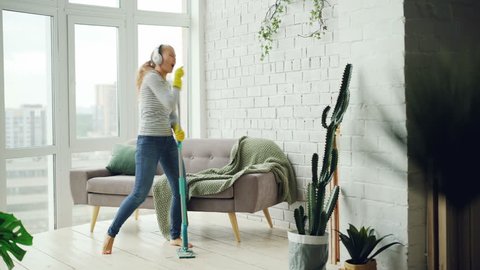 Joyful girl is mopping floor in light apartment and having fun listening to music through headphones, dancing and singing. Beautiful furniture and plants is visible.