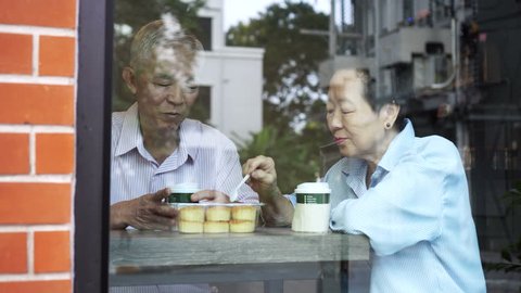 Happy Asian elderly senior couple drinking morning coffee through cafe glass front