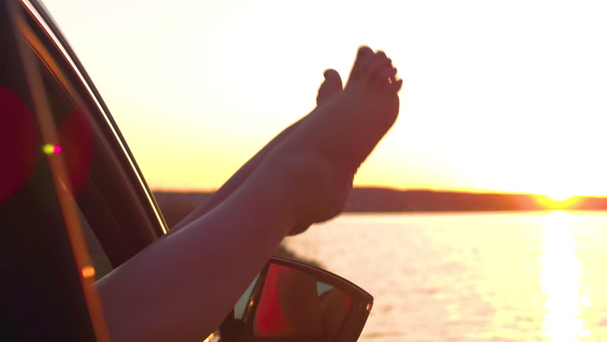 CLOSE UP, LENS FLARE, COPY SPACE: Unrecognizable carefree girl kicking her legs up and down through the car window on a beautiful summer evening by the beach. Cheerful woman enjoying a scenic sunset.