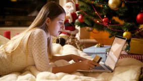 4k video of beautiful smiling young woman lying under Christmas tree and ordering gifts at online shop on laptop