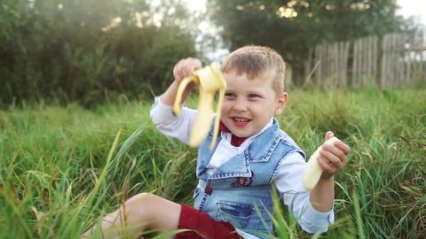 A little boy catches a peel from bananas. Cute baby eating banana and smiling. Handsome kid with a banana in his hand. The child laughs at the camera and eats fruit. The boy in nature on a picnic.