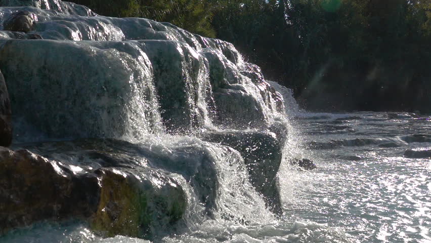 SLOW MOTION: Hot thermal water flowing down rocks splashes and glimmers in the bright sunlight. Glassy natural spring water flows from one geothermal pool to another on a sunny day in scenic Tuscany.