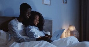 Portrait shot of the happy African American couple sitting with laptop computer and hugging in the bed late in the evening.