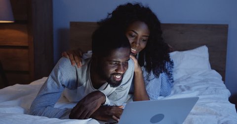 Portrait shot of the young African American couple lying on the bed at night, watching something on the laptop computer and then looking at each other.