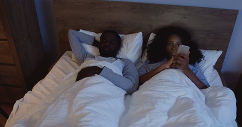 African American young woman lying in the bed and using her smartphone at night, her boyfriend waking up and talking to her, then embracing.
