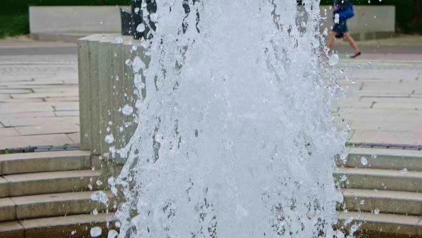 Urban rejuvenation: striking fountain erupts with forceful spray, generating dynamic eruption of crystalline droplets that shimmer in ambient illumination. Modern architectural setting features smooth, geometrically arranged stone steps descending to paved plaza, creating structural contrast with fluid aquatic display. Ambient figures in casual summer garments stroll through scene, one wearing blue shirt with shorts, evoking warm inviting atmosphere. Natural diffused lighting brightens cheerful environment, emphasizing vibrant aquatic textures and reflections on polished stone surface. Dynamic composition captures essence of metropolitan leisure and public enjoyment with lively atmosphere perfect for urban planning concepts. Refreshing display conveys sense of community revitalization, ideal for tourism promotions or social content about city parks.