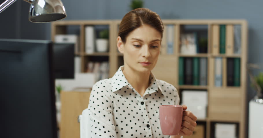 Close up of the young beautiful woman sitting at the computer in her office and drinking coffee or tea.