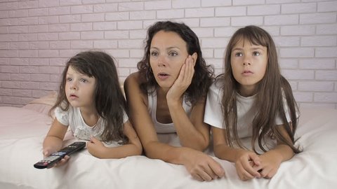 The family is watching TV. Children with their mother in bed watching TV.