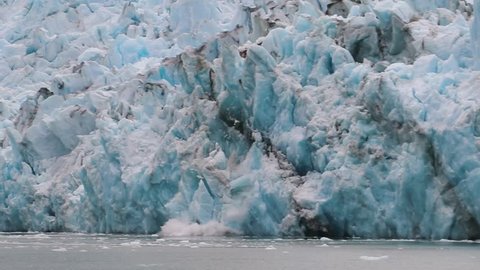 Slow motion, medium wide view of Dawes Glacier calving. Large chunks of jagged blue ice break from the face of the glacier and fall into the cold water of the fjord to form icebergs. Stock Video