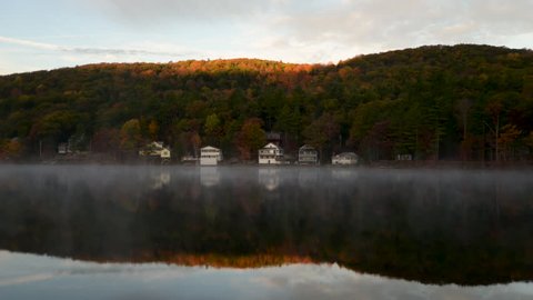 Timelapse morning fog drifts across a still lake surrounded by colorful trees in autumn during a sunrise.