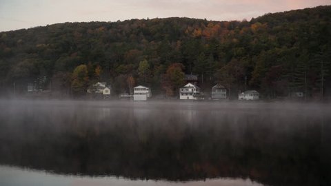 Morning fog drifts across a still lake surrounded by colorful trees in autumn.