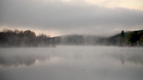 Morning fog drifts across a still lake surrounded by colorful trees in autumn.
