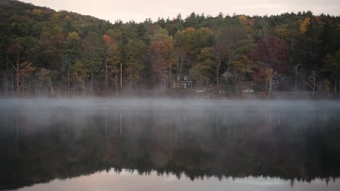Morning fog drifts across a still lake surrounded by colorful trees in autumn as a car drives by.