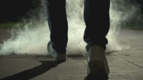 The legs of a man walking near a cloud of smoke. evening night time, slow motion