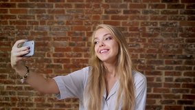 Young caucasian blonde making selfie, smiling, photo, smartphone, portrait, brick wall in the background.