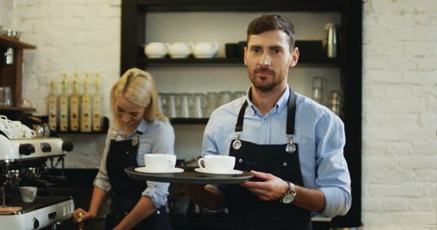 Portrait shot of the handsome Caucasian barmen holding cups of coffee and going to the bar while his female co-worker preparing more behind at the coffee machine.