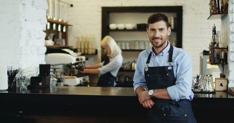 Portrait shot of the handsome smiled waiter standing and leaning on the bar while blond attractive waitress preparing coffe behind.