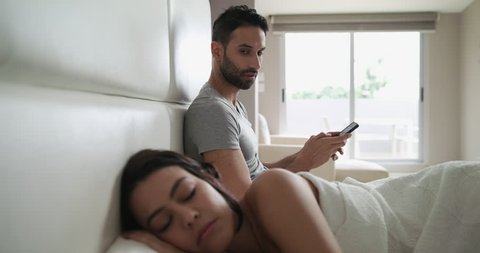 Young latino man and woman in bed at home. Hispanic husband texting with smartphone and sending message to his lover while his wife is still sleeping