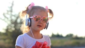 Funny child in headphones listens to music, sings and dances. Lovely girl in sunglasses at sunset on a summer day.