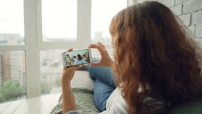 Girl with curly hair is making video call chatting to friends sending hand kisses and waving hand lying on sofa at home. Brick wall and panoramic windows are visible.