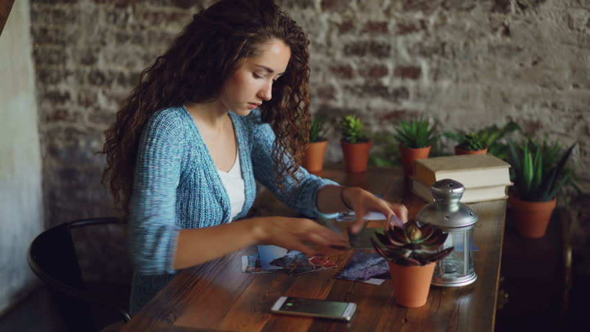 Pretty girl creative designer is putting photographs on wooden table then shooting flat lay pictures with smartphone. Modern technology, illustrations and people concept.