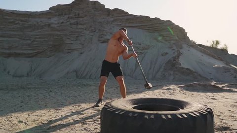 Muscle athlete strongman man hits a hammer on a huge wheel in the sandy mountains in slow motion. strength and endurance training for wrestlers