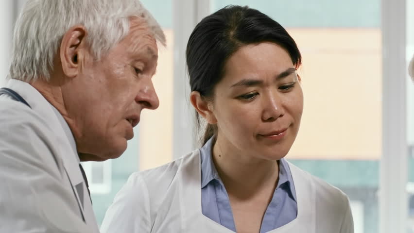 Tilt up of senior male doctor explaining x-ray results to Asian female colleague while working in team in medical clinic | Shutterstock HD Video #1016037136