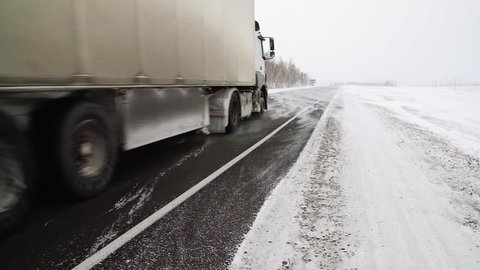 The car is on a snow-covered highway.
Heavy truck with trailer carries cargo. 
Winter road with a gusty wind and a snowstorm, slow motion.