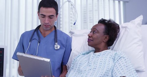 Young hispanic doctor reviewing health history with senior African patient. Middle aged black woman lying in hospital lstening to charming young MD explain her current health condition
