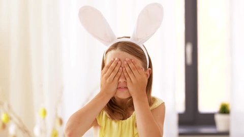 easter, holidays and people concept - happy girl wearing bunny ears headband playing peek a boo game at home