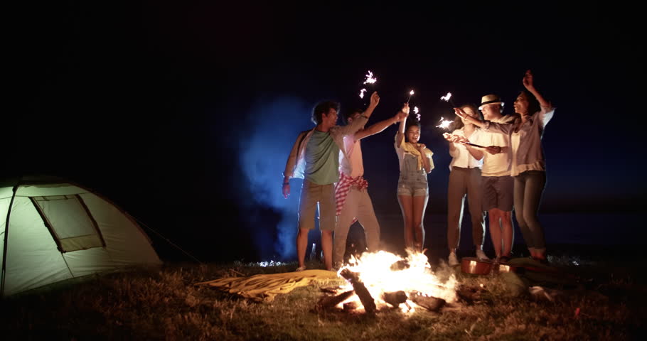 Six friends dancing near campfire during summer party, smiling, cheering, holding sparklers in hands, enjoying their time 4k