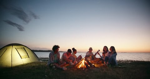 Group of six old friends having a summer beach party near bonfire, talking, smiling, drinking beer, wine, toasting and cheering having fun 4k 스톡 비디오