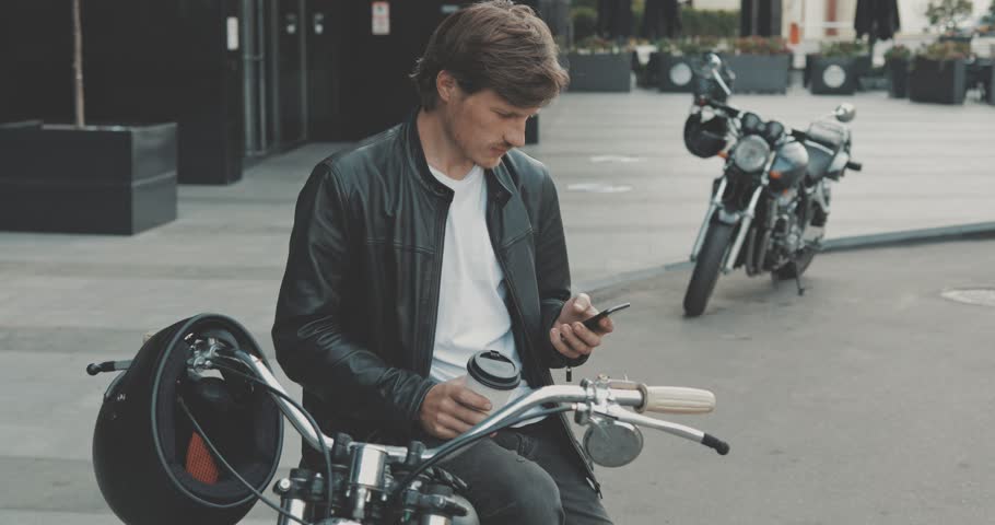 Guy biker drinking coffee while standing near classic motorcycle from 1970s on street and writing message on smartphone phone. Urban lifestyle scene. 4K video shooting by handheld gimbal