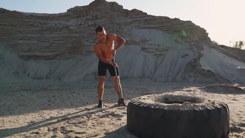 Muscle athlete strongman man hits a hammer on a huge wheel in the sandy mountains in slow motion at sunset. The dust from the wheels rises.