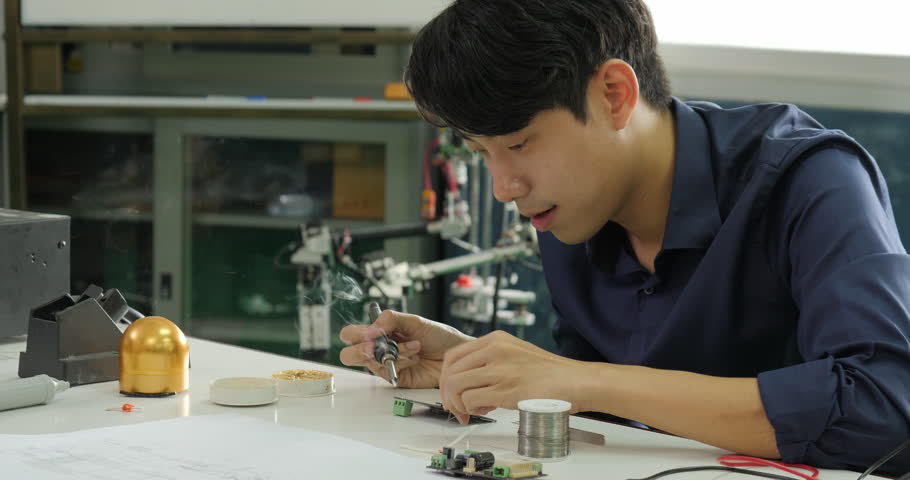 Asian electronics engineer is soldering an electric board in his workshop. Asian student are studying electronics and soldering in classroom.